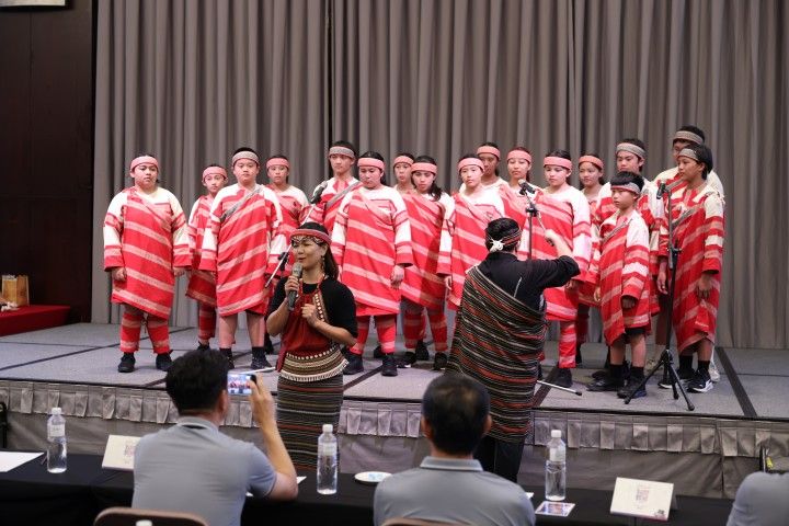 合唱團登台｜紅白條紋族服的孩子站滿舞台，領唱在前，指揮背對著觀眾張開雙手。